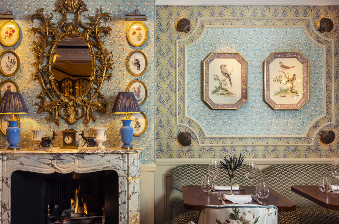 A table for two next beside the antique fireplace. Walls feature cream decorative panelling, pastel wallpapers from Fromental, shell-shaped lights and colourful illustrations in gold frames. A carriage clock, fawn figurines, a white vase and a pair of blue lamps are on the mantlepiece